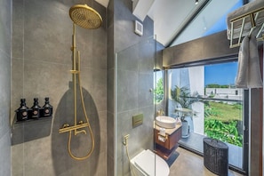 Un baño moderno con cabina de ducha de vidrio, inodoro y lavamanos con vistas a la vegetación exterior.