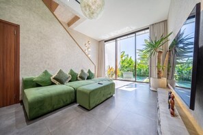 Ein modernes Wohnzimmer mit einem Ecksofa, einem Flachbildfernseher und einem großen Fenster mit Blick auf Grünflächen.
