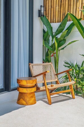 A wicker chair with a wooden base and a round table beside it, positioned on a balcony with a white wall and bamboo backdrop. - Tropical 2BR Gem in Ubud w Private Pool & Gazebo (Gianyar Regency)