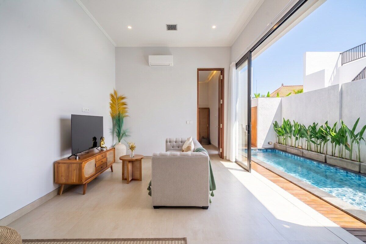 A modern living room with a flat-screen TV, a sofa, a small side table, and a poolside area with a view of the outside.