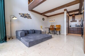 A modern living room with a sectional sofa, a dining table with chairs, a floor lamp, and a kitchen area visible through an open doorway. - Cozy 1BR Gem in Ubud w Pool • Perfect for Couples! (Kabupaten Gianyar)