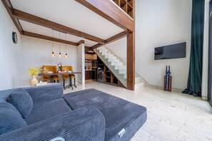 A modern living room with a sofa, dining area, staircase, and a flat-screen TV. - Cozy 1BR Gem in Ubud w Pool • Perfect for Couples! (Kabupaten Gianyar)