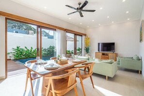 A modern living room with a dining area, a television, and a large window overlooking a garden. - Chic 3BR Villa 300m to Sanur Beach • Pool & Patio (Kota Denpasar)