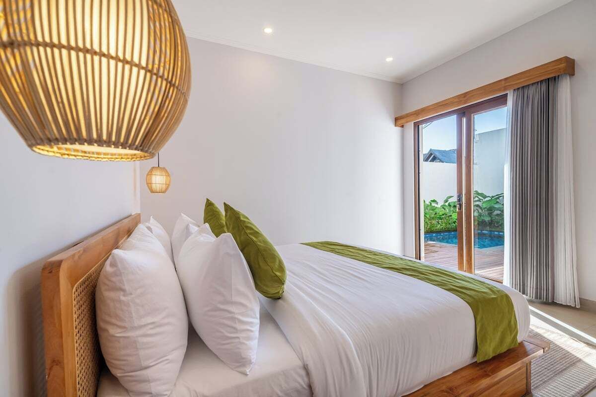 A bedroom with a bed, pillows, a hanging light fixture, and a view of greenery outside.
