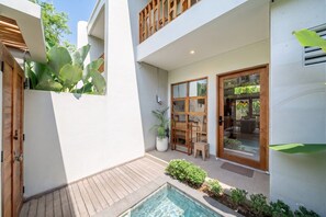Un espacio exterior moderno con terraza de madera, una piscina pequeña y un ventanal que da a un área interior con mobiliario.