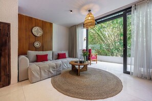 A modern living room with a sofa, a coffee table, and a large window with a view of greenery. - Cozy 1BR Flat in Ubud • Pool • Kitchen • Garden (Gianyar Regency)