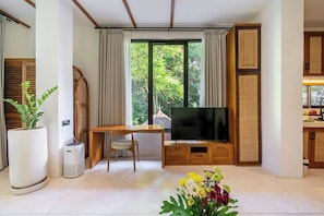 A modern living room with a large window, a flat-screen TV, a wooden desk, a chair, and a potted plant. - Stylish 1BR in Ubud w Pool, Patio, Private Kitchen (Gianyar Regency)