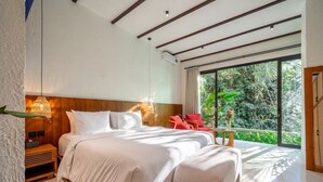 A modern bedroom with a large bed, a wooden headboard, a bedside table, a red chair, and a large window with a view of greenery. - Tropical 1BR Stay in Ubud w Pool, Patio & Kitchen (Gianyar Regency)