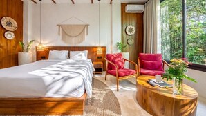A modern bedroom with a large bed, a red chair, a round coffee table, and a view of greenery through the window. - Best 1BR Stay in Ubud • Pool • Garden • Kitchen (Gianyar Regency)