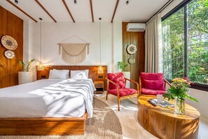 Quarto moderno com cama grande, cadeira vermelha, mesa de centro redonda e vista para o verde pela janela.