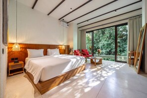 A modern hotel room with a large bed, a small table, a chair, and a large window offering a view of greenery. - Designer 1BR Flat in Ubud w Pool, Garden & Kitchen (Gianyar Regency)