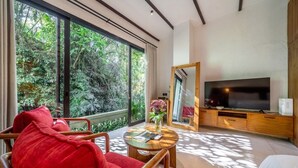 A modern living room with a large window, a flat-screen TV, and a cozy seating area. - Designer 1BR Flat in Ubud w Pool, Garden & Kitchen (Gianyar Regency)