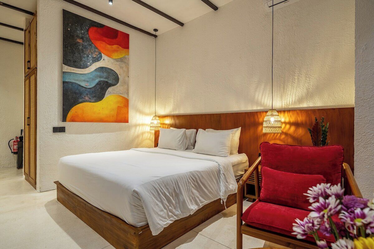 A bedroom with a bed, a red chair, a wall painting, and a wall-mounted lamp.