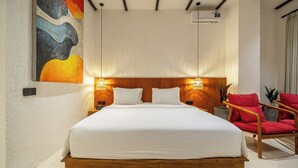 A bedroom with a wooden bed frame, white bedding, two bedside lamps, a red chair, and a wall-mounted air conditioning unit. - Designer 1BR Flat in Ubud w Pool, Garden & Kitchen (Gianyar Regency)