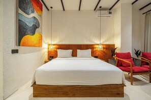A bedroom with a wooden bed frame, white bedding, two bedside lamps, a red chair, and a wall-mounted air conditioning unit. - Designer 1BR Flat in Ubud w Pool, Garden & Kitchen (Gianyar Regency)