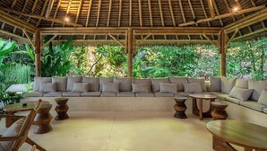 A covered outdoor seating area with a thatched roof, cushioned sofas, and wooden tables. - Designer 1BR Flat in Ubud w Pool, Garden & Kitchen (Gianyar Regency)