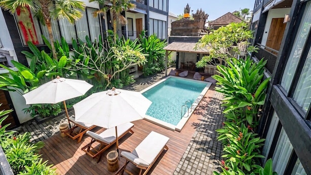 Uma área moderna com piscina ao ar livre, espreguiçadeiras e guarda-sóis, cercada por vegetação exuberante e um templo balinês tradicional ao fundo.