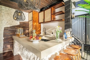 A kitchen with a stone wall, wooden cabinets, a fruit bowl, and a bar area with stools. - Chic 1BR • Pool • Private Kitchen • Central Ubud (Kabupaten Gianyar)