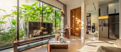 Un salon moderne avec un grand téléviseur, un canapé et une table basse. Une grande fenêtre donne sur l’extérieur, où l’on peut voir des plantes.