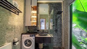 Un baño moderno con lavadora integrada, ducha envuelta en vidrio y un amplio ventanal con vistas a zonas verdes.