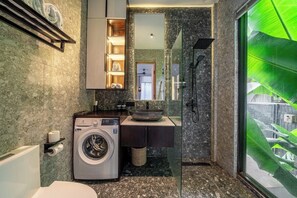 Ett modernt badrum med en inbyggd tvättmaskin, en dusch med glasväggar och ett stort fönster med utsikt över grönområden.