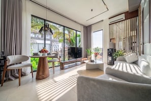A modern living room with a flat-screen TV, a sofa, a dining table with chairs, and a large window with a view of palm trees. - Hidden 1BR Gem in Central Ubud w Pool & Kitchen (Kabupaten Gianyar)