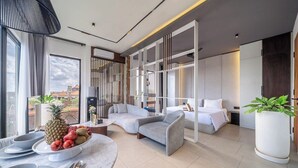 Una habitación de hotel moderna con un sofá, una mesa de comedor con fruta, una planta grande en maceta y vistas a la ciudad.