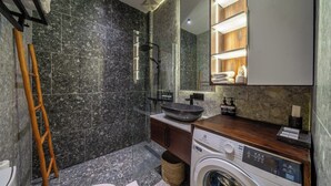 Un baño moderno con pared de mármol, cabina de ducha de vidrio, tocador de madera y lavadora integrada.