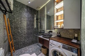A modern bathroom with a marble wall, a glass shower enclosure, a wooden vanity, and a built-in washing machine. - Serene 1BR Stay in Ubud w Pool & Private Kitchen (Ubud)