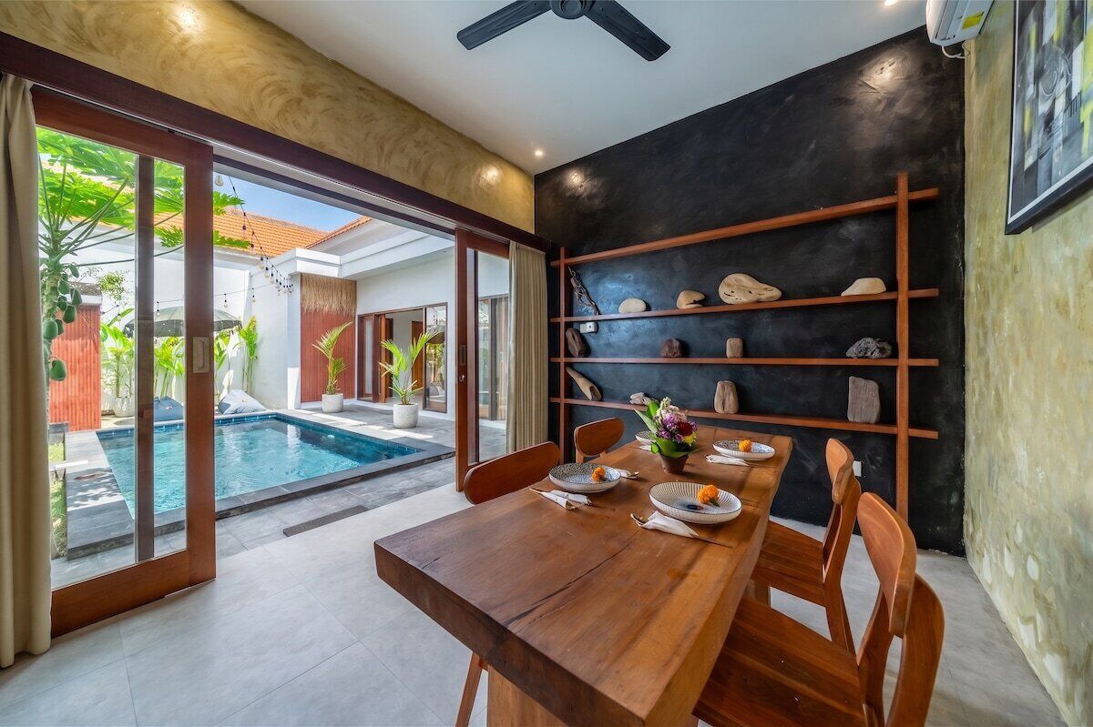 A dining area with a wooden table and chairs, a black wall with shelves, and a view of a swimming pool.