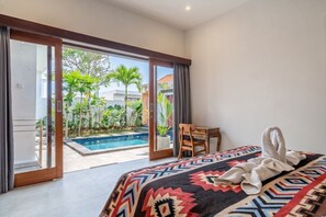 A bedroom with a bed, a desk, and a view of a swimming pool and greenery. - Tropical 2BR Villa w Beach Nearby, Patio & Pool (Mengwi)