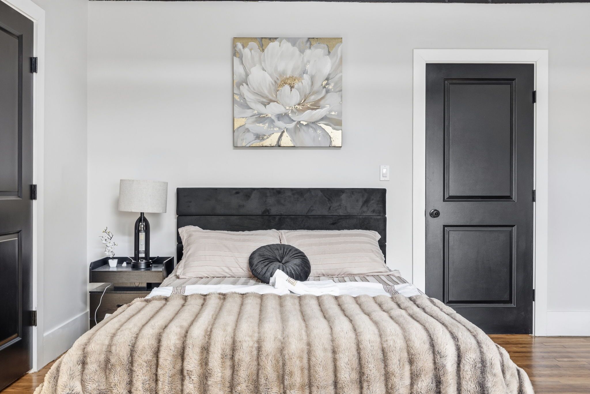 Un dormitorio con cama, mesita de noche, lámpara, puerta y una obra de arte en la pared.