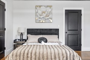 A bedroom with a bed, a nightstand, a lamp, a door, and a wall art piece. - Bright and spacious 3BR Apt, gated 2 car parking, close to T station (Boston)