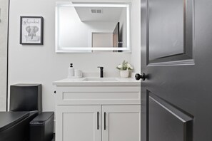 Une salle de bain moderne avec un meuble-lavabo blanc, un grand miroir, un robinet noir et un tableau encadré au mur.