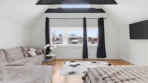 A loft bedroom with a sloped ceiling, a large window with curtains, a sofa, a coffee table, and a rug.