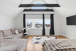 Une chambre à coucher aménagée dans un espace mansardé, avec un plafond en pente, une grande fenêtre avec des rideaux, un canapé, une table basse et un tapis.