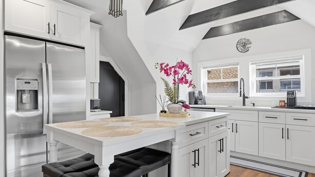 A modern kitchen with white cabinetry, a stainless steel refrigerator, a water dispenser, and a dining area with a bench.