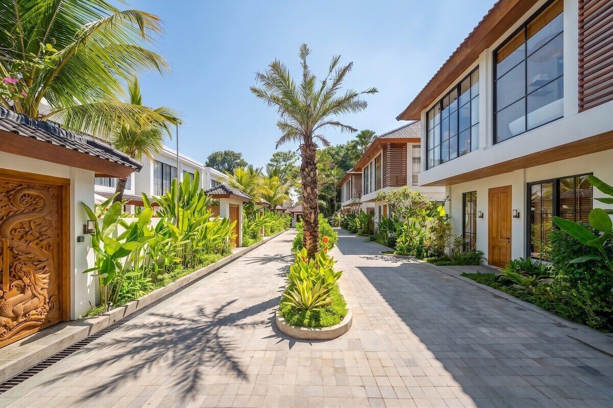A pathway flanked by modern buildings with large windows, surrounded by lush tropical plants.