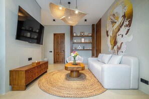 A modern living room with a flat-screen TV, a wooden console, a white sofa, a woven rug, and a decorative wall piece. - Serene 1BR Stay in Central Ubud w Pool & Kitchen (Gianyar Regency)