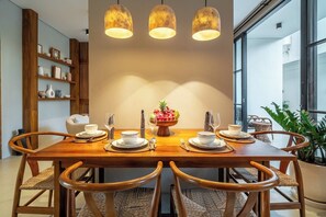 Uma área de refeições com mesa de madeira posta para quatro pessoas, rodeada por cadeiras de vime, um centro de mesa com tigela de frutas e iluminação pendente.