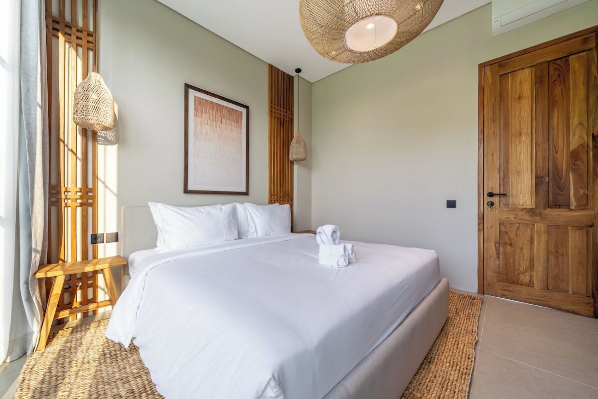 Cama bem arrumada com lençóis brancos, porta de madeira e tapete de palha.
