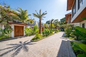 A pathway flanked by lush greenery and palm trees, leading to a building with a traditional tiled roof. - Cozy 1BR • Ubud • Pool • Private Kitchen • Garden (Gianyar Regency)