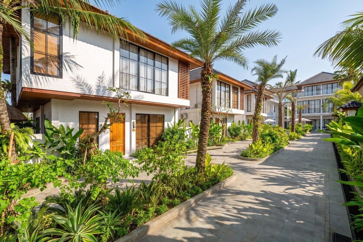 A row of modern villas with large windows, surrounded by lush tropical plants and a paved walkway.