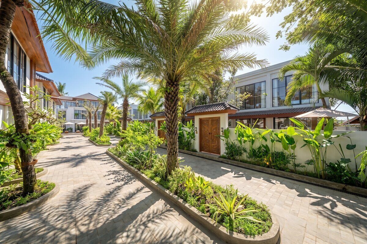 Un camino bordeado de palmeras y plantas tropicales que conduce a un edificio con tejado de baldosas tradicionales.