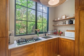 A kitchen with wooden cabinets, a gas stove, a sink, and a large window offering a view of greenery. - Serene 1BR • Ubud • Pool • Private Kitchen & Patio (Gianyar Regency)