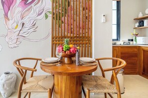 A dining area with a round wooden table, two chairs, a fruit bowl, and a decorative wall panel. - Luxe 1BR in Ubud w Pool, Private Kitchen & Balcony (Gianyar Regency)