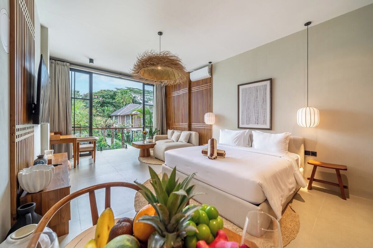 A hotel room with a bed, a dining table with fruits, a sofa, and a balcony with a view of greenery.