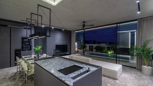 A modern kitchen with a central island, dining area, and a large window overlooking a pool. - Designer 2BR Villa in Mengwi w Pool & Beach Nearby (Kabupaten Tabanan)