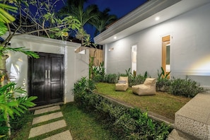Une maison moderne avec une porte sombre, un petit jardin avec de la pelouse et des plantes, et deux chaises de couleur beige.