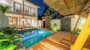 Een modern huis met twee verdiepingen, een zwembad, een houten terras en een rieten dakconstructie.
